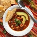 Serving bowl and spoon with chicken tortilla soup showing hominy, black beans, and corn. It's topped with cilantro, 2 slices of avocado, with 3 tortilla chips are tucked on the side.