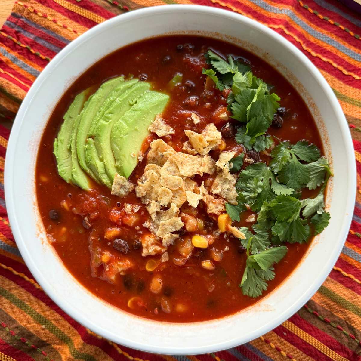 Large serving bowl with chicken tortilla soup with hominy, black beans, corn topped with crushed tortillas, cilantro, and slices of avocado.