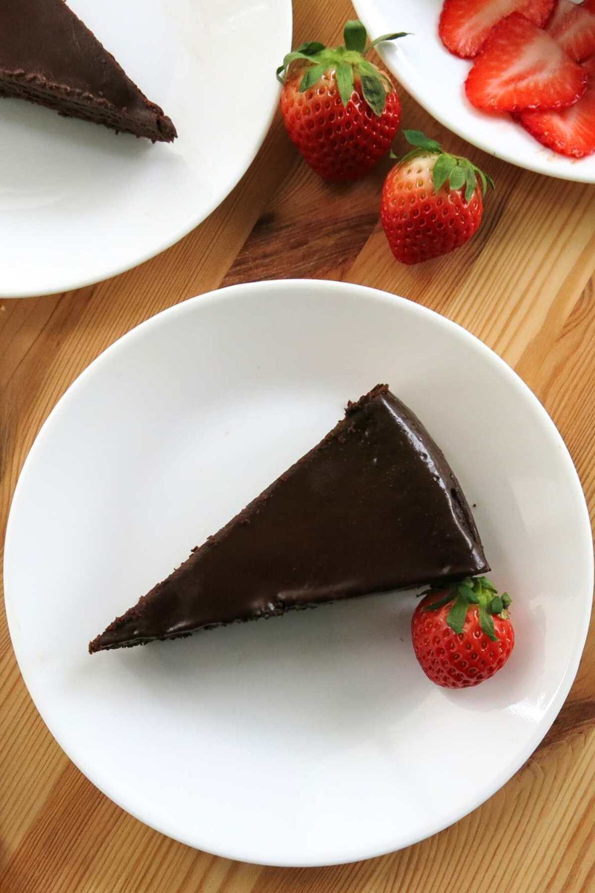 Slice of flourless chocolate cake covered with ganache on a white plate with a strawberry with more strawberries and cake behind it.