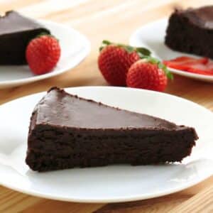 Slice of flourless chocolate cake with ganache on a white plate with strawberries and more plates with cake behind it.