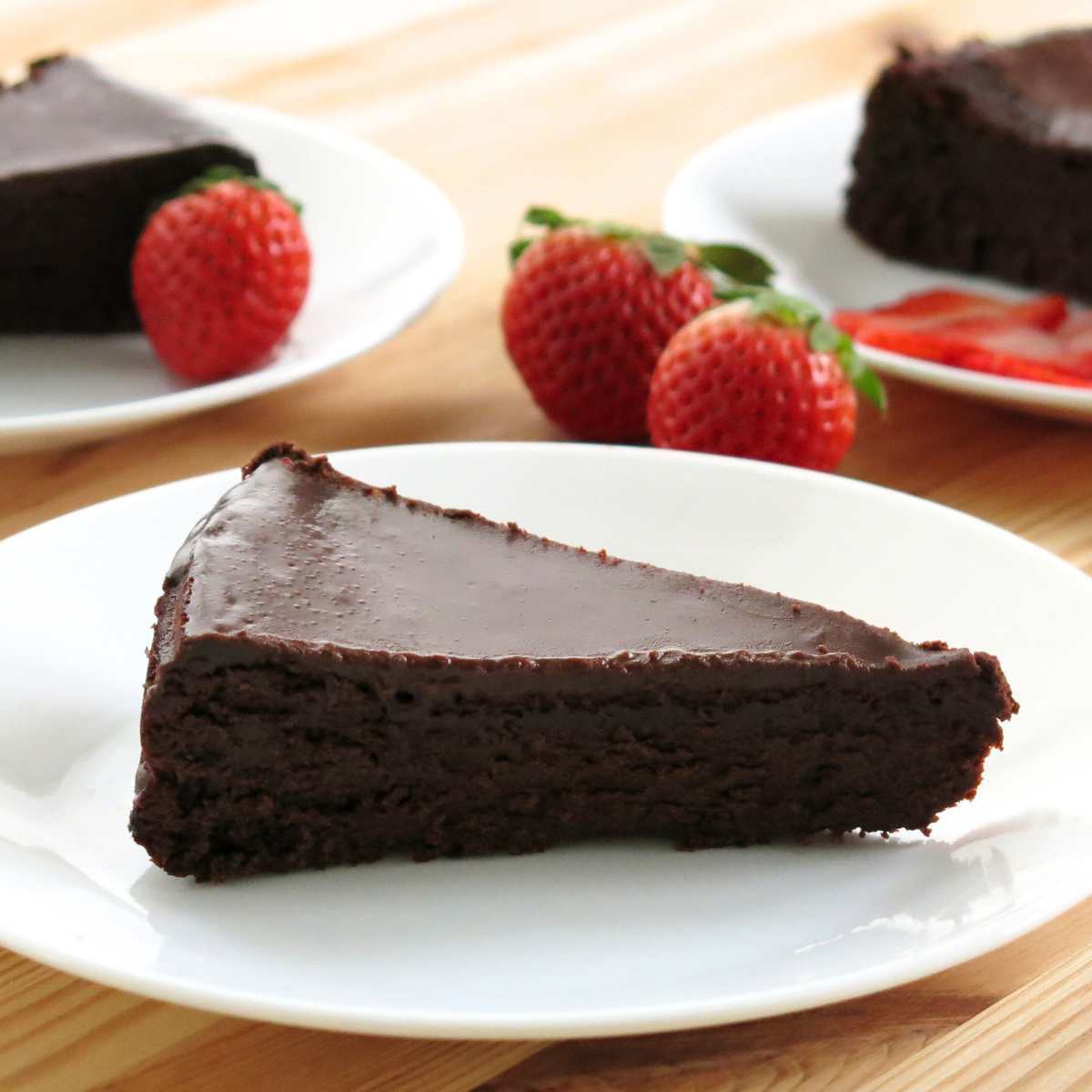 Slice of flourless chocolate cake with ganache on a white plate with strawberries and more plates with cake behind it.