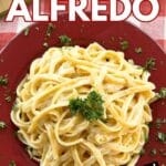 Bowl of fettuccine alfredo in a red bowl with Parmesan cheese and parlesy behind it.