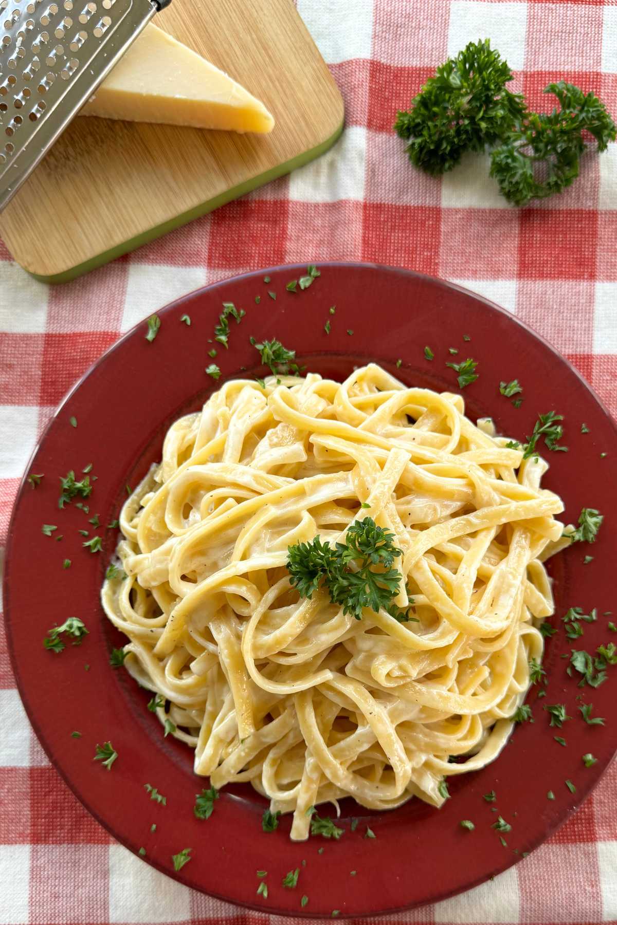 Creamy fettuccine alfredo in a red bowl garnished with parsley with a triangle of Parmesan cheese and a bunch of parsley behind it.