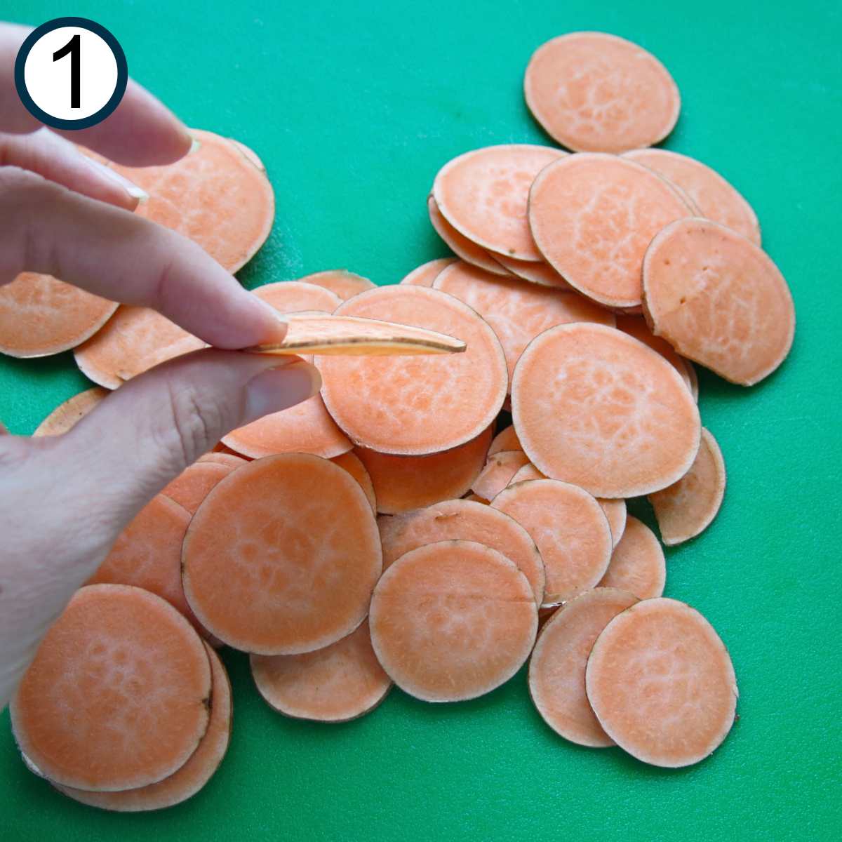 Fingers holding a thin slice of a sweet potato over a pile of more on a cutting board.