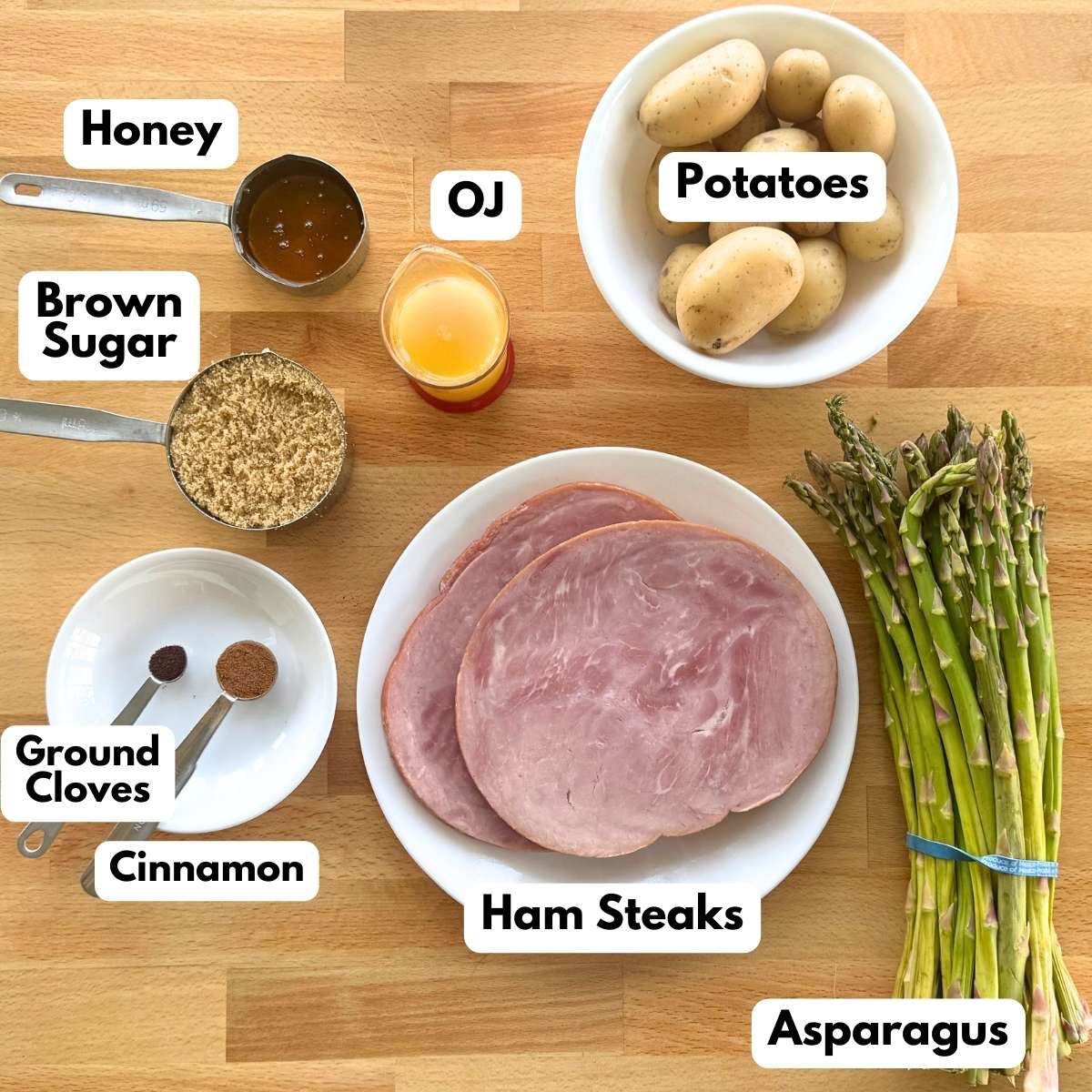 Two ham steaks, honey, orange juice, brown sugar, cinnamon, ground cloves, asparagus, and small potatoes on a cutting board.