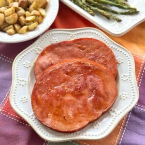 Two baked ham steaks coated with a honey, orange juice, brown sugar glaze on a white plate with asparagus and roasted potatoes behind it.