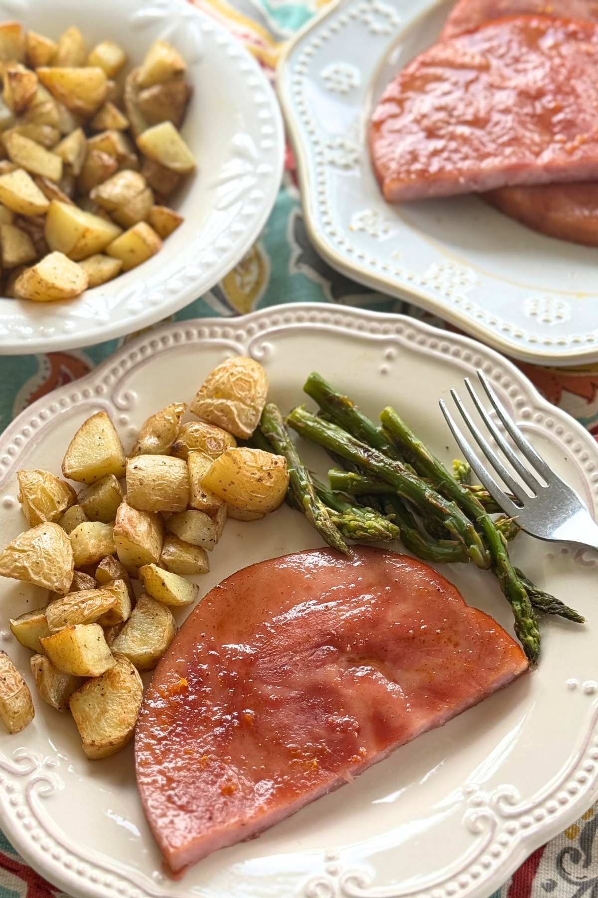 Serving of ham steak, roasted potatoes, and asparagus on a plate with more potatoes and slices of ham in the background.