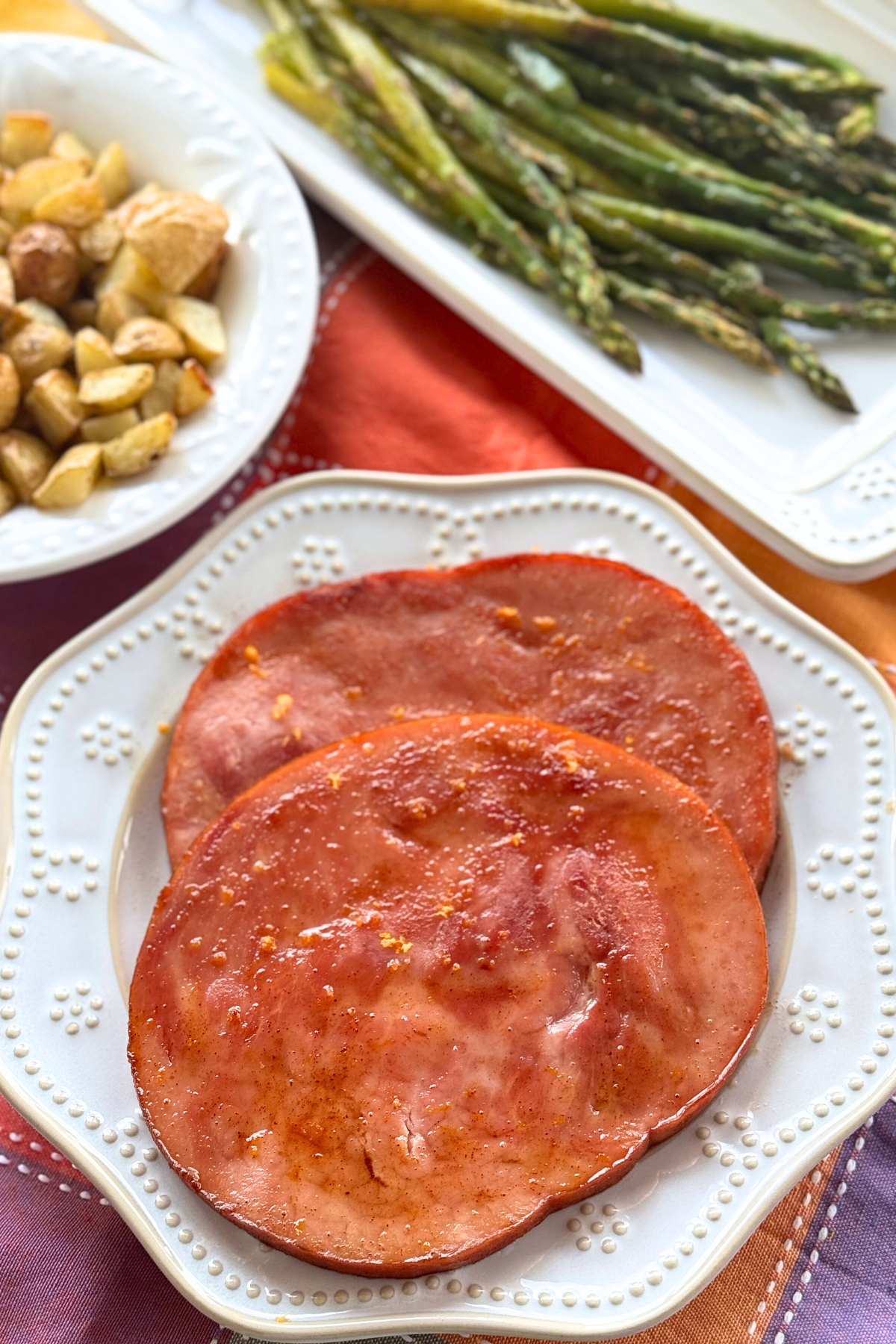 Two glazed ham steaks on a plate with asparagus and roasted potatoes in dishes behind it.