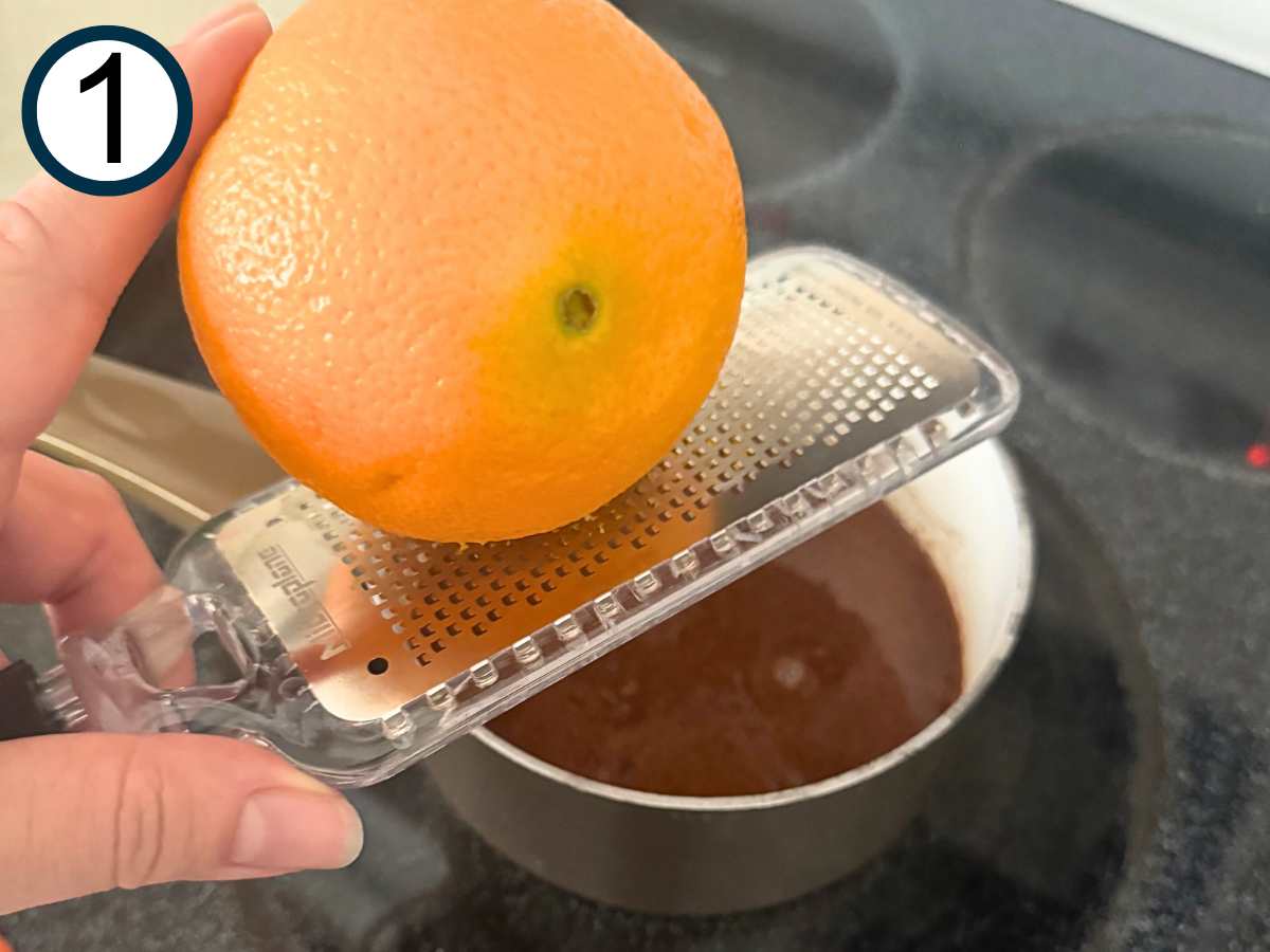 Orange being zested over a small sauce pot with a brown mixture of brown sugar, orange juice, honey, and spices.