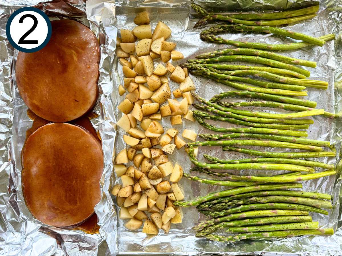 Uncooked ham steaks brushed with a honey orange glaze, diced pototes, and asparagus spears on a large sheet pan lined with aluminum foil.