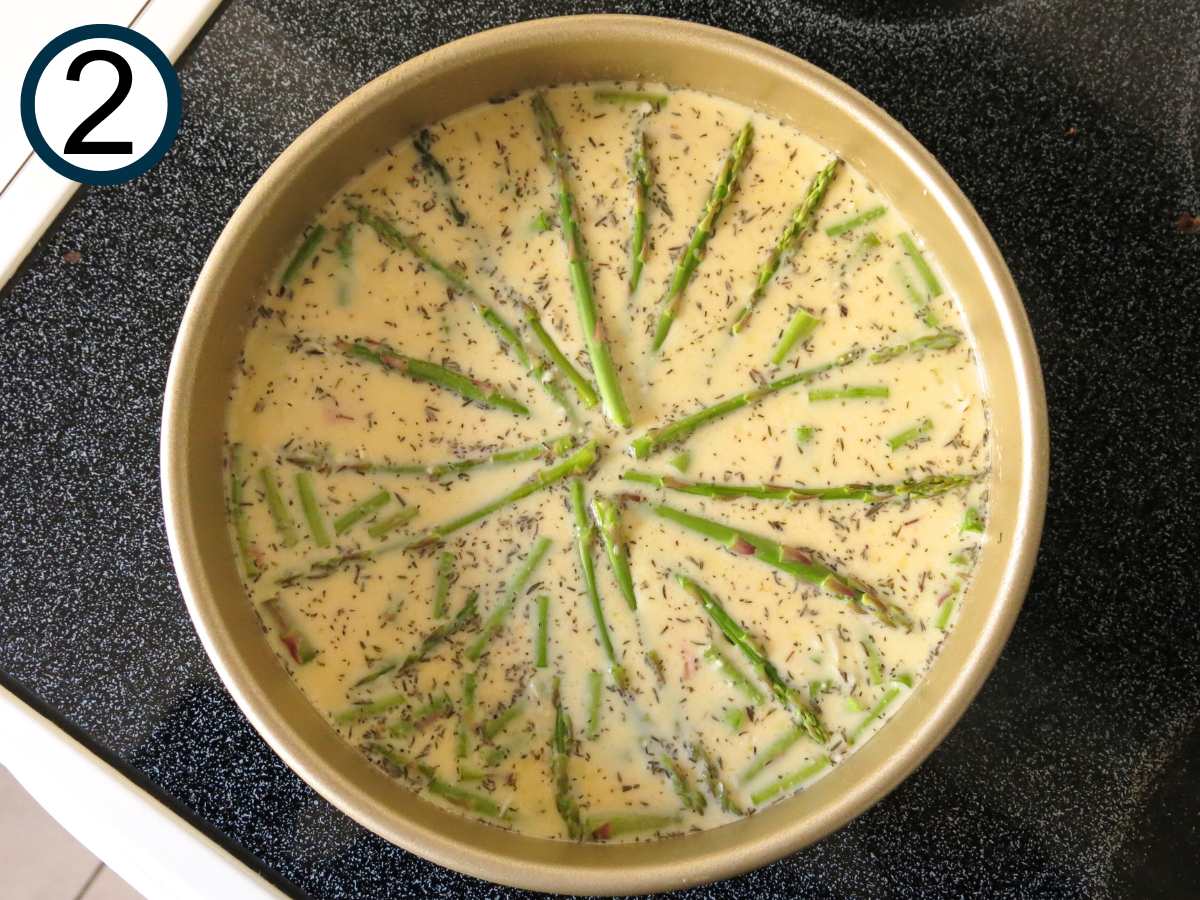 Uncooked quiche in a round cake pan with asparagus spears arranged in a sunburst pattern.