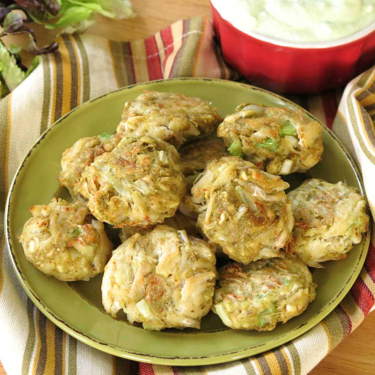 Avocado crab cakes on a plate with a bowl avocado crema and lettuce behind it.