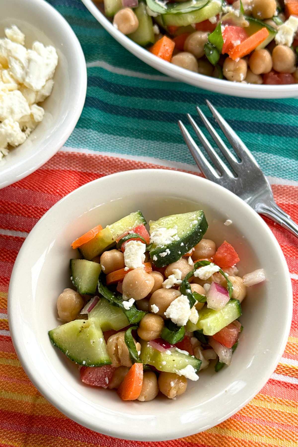 Small bowl with chickpea cucumber feta salad next to a fork with a bowl of crumbled feta cheese and larger bowl of salad behind it.