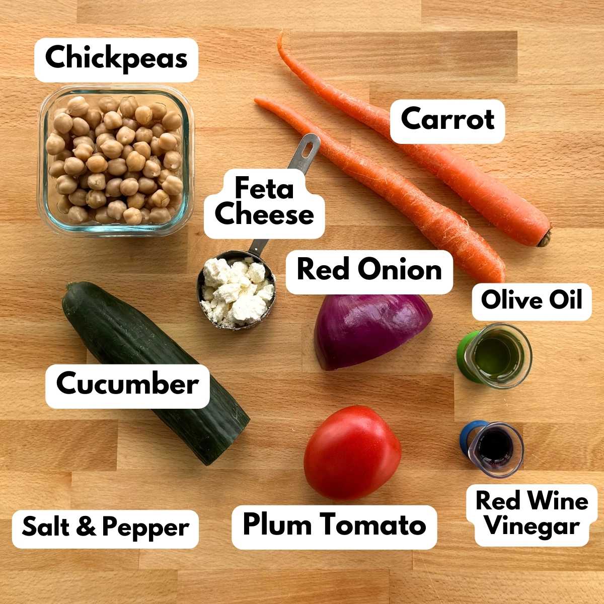 Labeled ingredients to make chickpea salad recipe: chickpeas, crumbled feta cheese, half an English cucumber, 2 carrots, plum tomato, red onion, olive oil, and red wine vinegar.