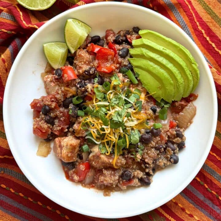 White bowl with a chicken, quinoa, black beans, and vegetables topped with shredded cheese and cilantro with avocado slices and lime wedges next to it.