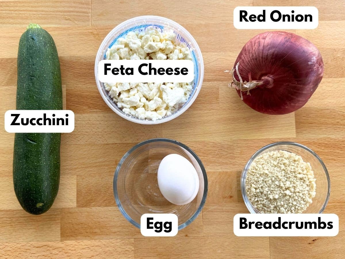 Ingredients to make the recipe: zucchini, crumbled feta cheese, red onion, egg, and panko breadcrumbs in a glass bowl.