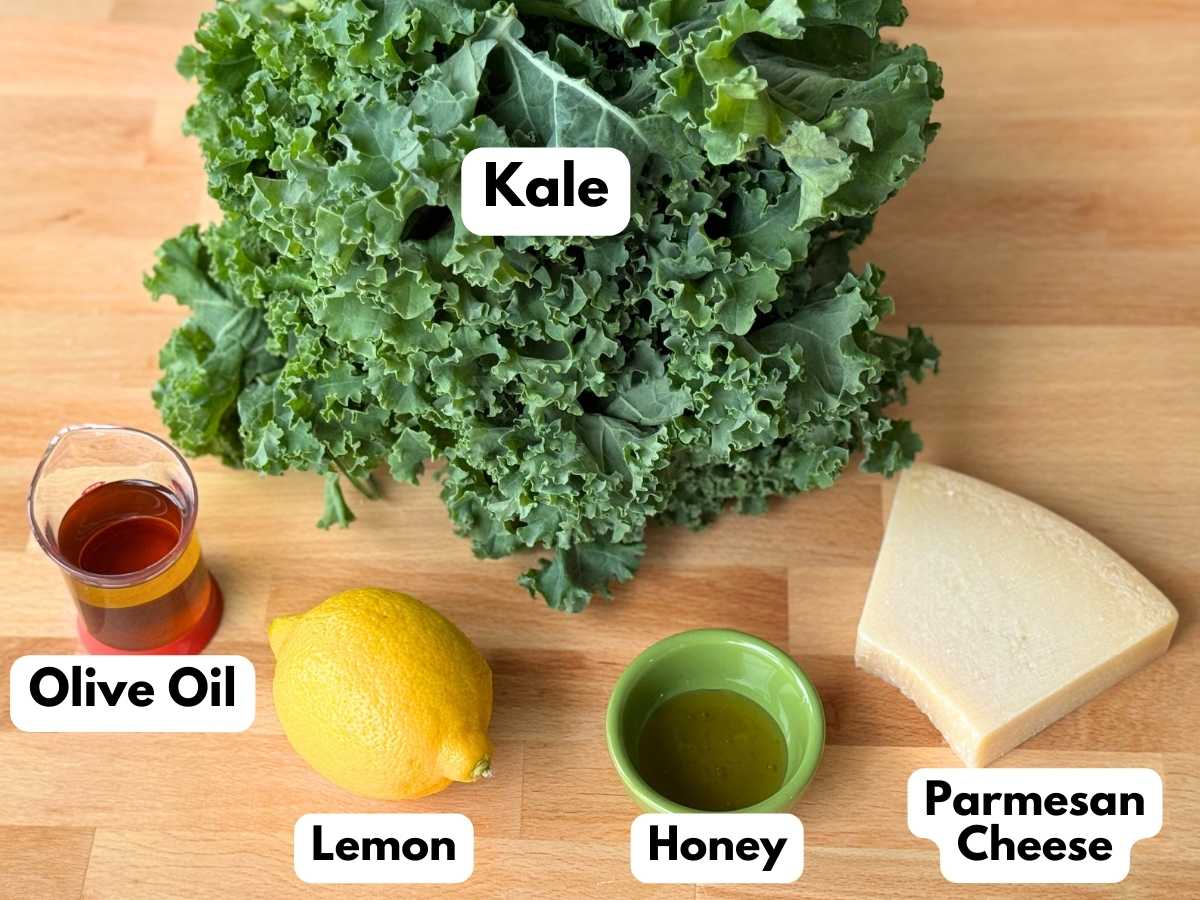 Ingredients to make recipe on butcher block background: bunch of curly kale, lemon, olive oil and honey cups, and a triangle of Parmesan cheese with some missing.