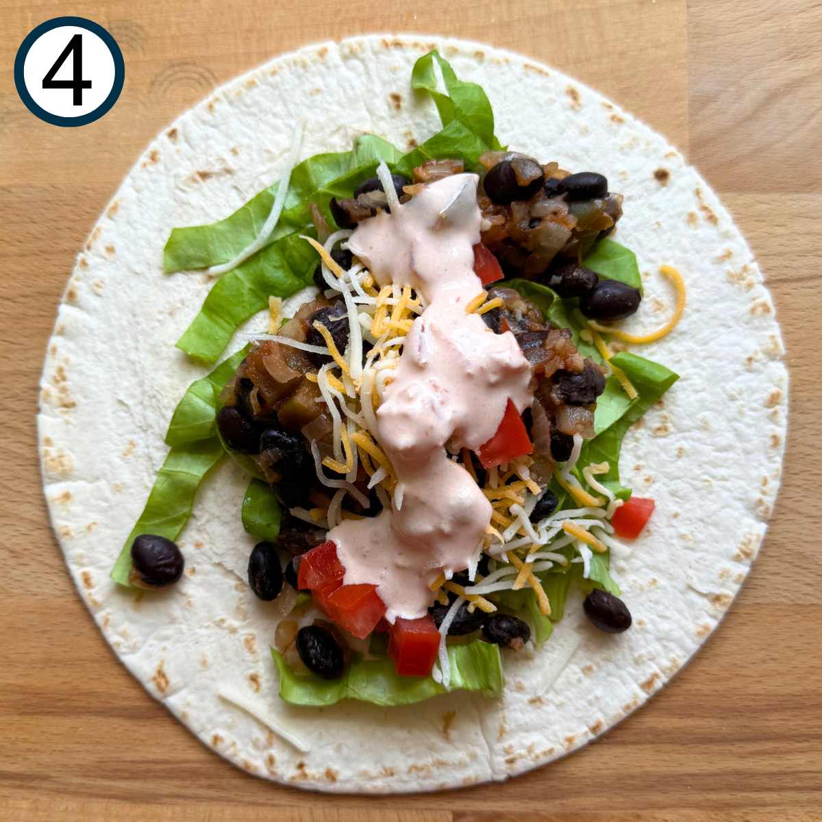 Flour tortilla on a cutting board with lettuce, black bean mixture, shredded cheese, and yogurt salsa sauce layered on top.