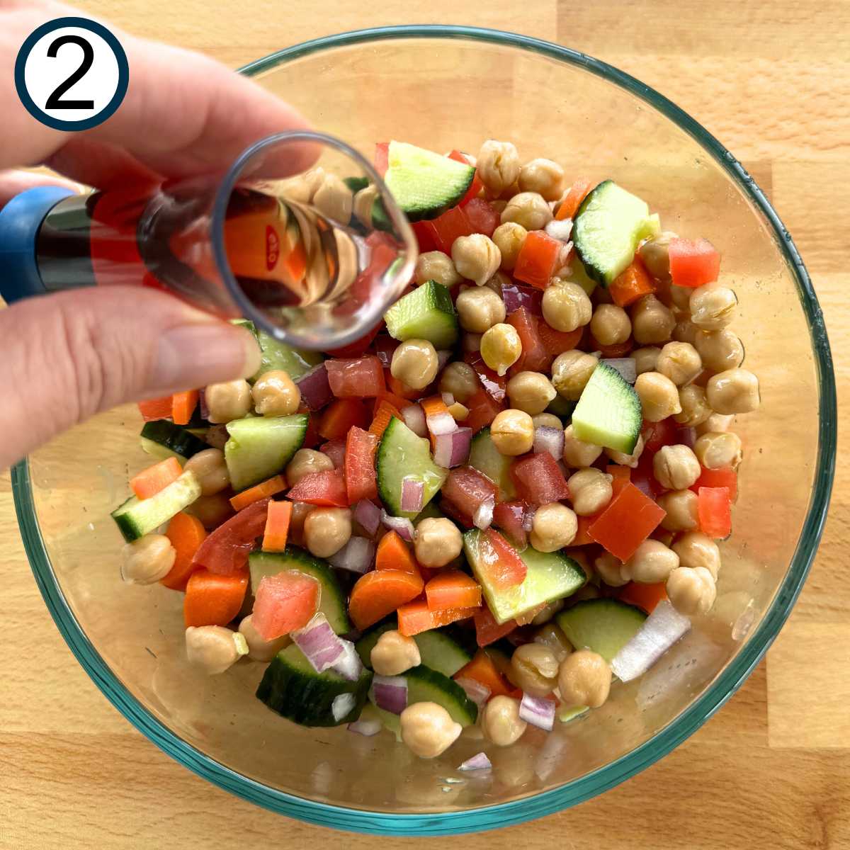 Hand pouring red wine vinegar over mixture of chickpeas, cucumber, tomatoes, red onion, and carrots.