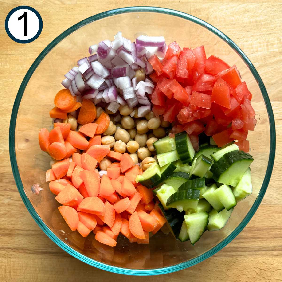 Glass bowl with chickpeas and diced cucumbers, carrots, tomatoes, and red onion.