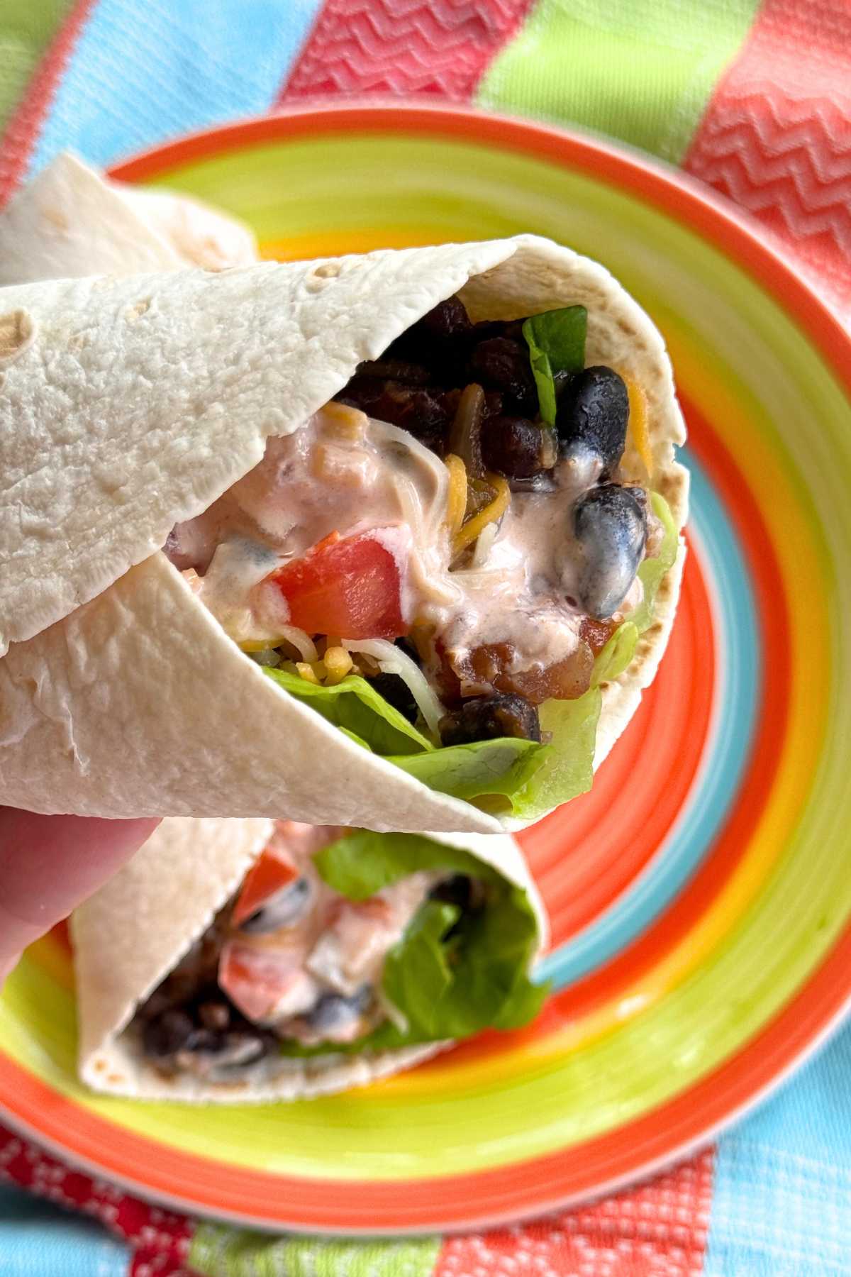 Hand holding a flour tortilla wrap with black bean, cheese, lettuce, tomato, and pink salsa and yogurt sauce over a colorful plate with another wrap. 