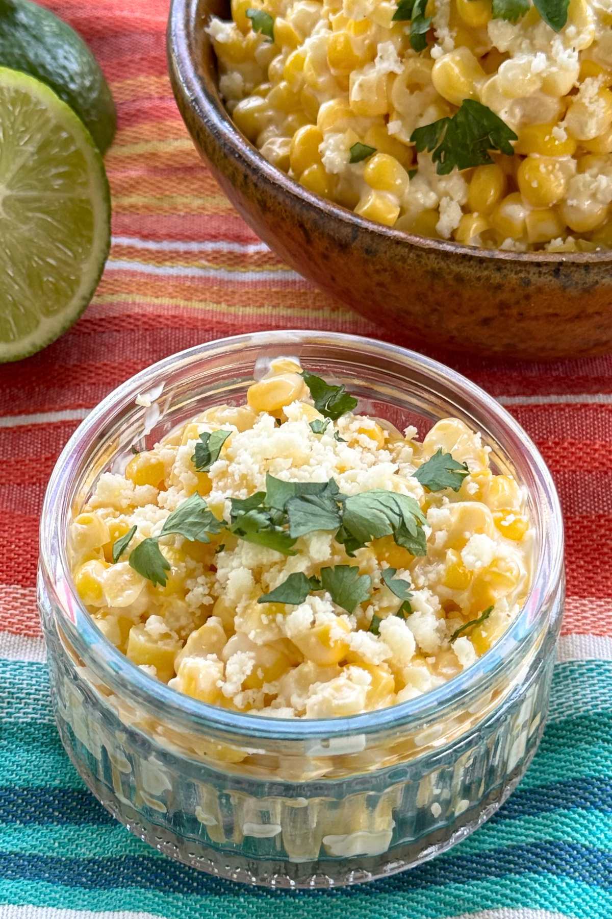 Mexican street corn in a glass cup with a lime and a bowl with more street corn behind it.