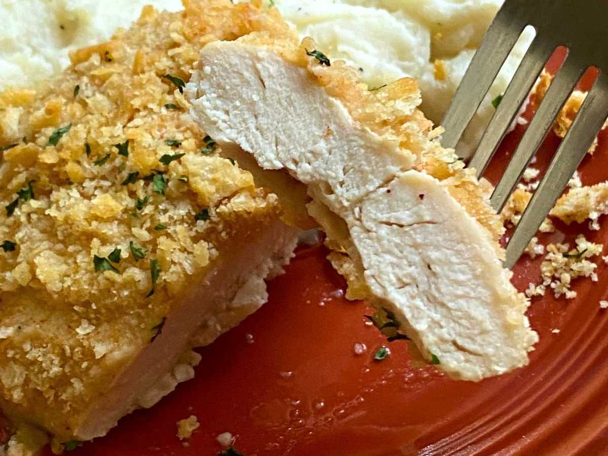 Fork holding a bite of heavenly chicken with the rest of the chicken breast coated with Ritz crackers on the plate with mashed cauliflower.