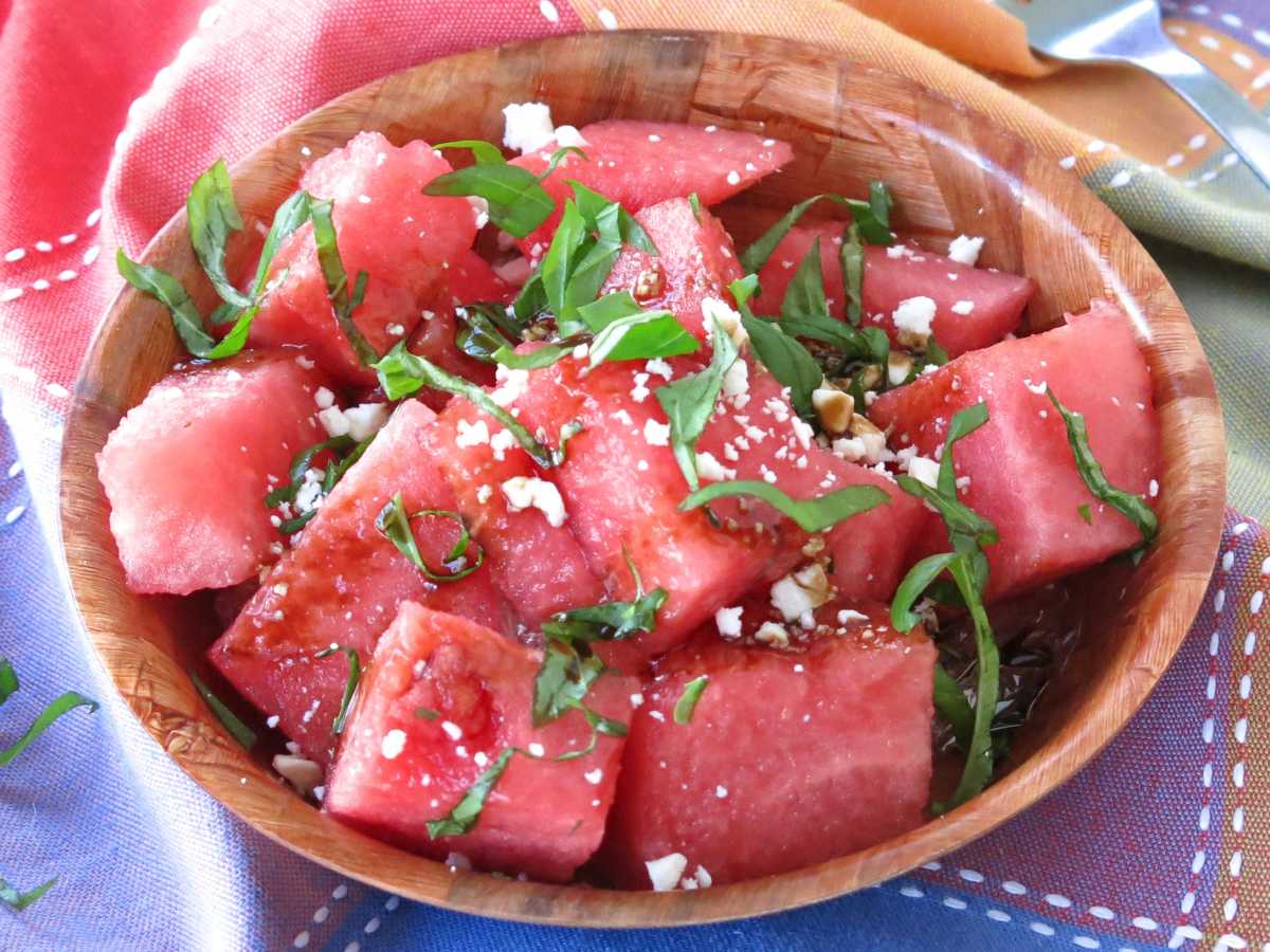 Chunks of watermelon topped with basil, balsamic glaze, and crumbled feta cheese in a brown bowl on a colorful napkin.