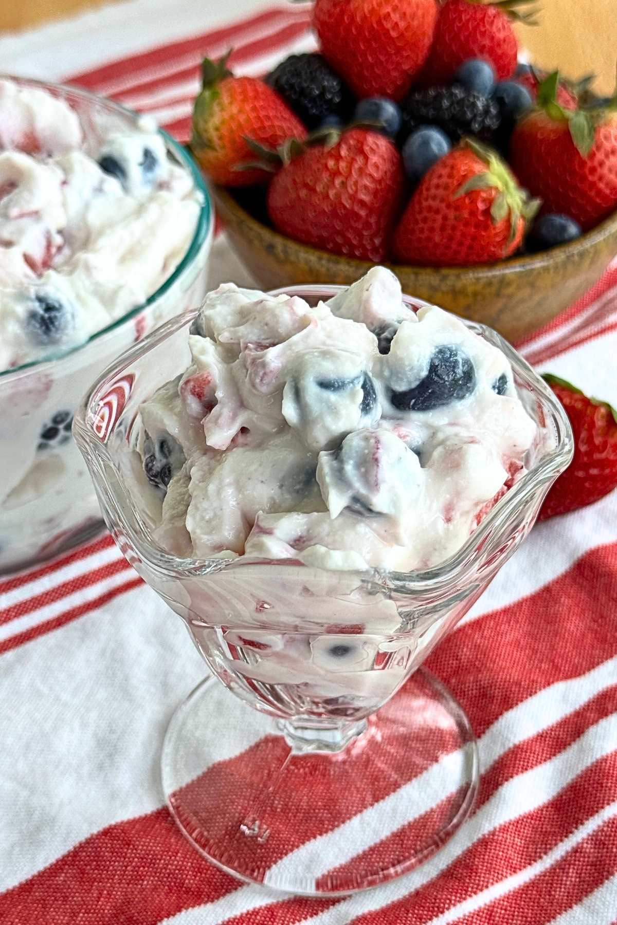 Glass sundae bowl filled with cottage cheesecake fruit salad with a bowl of fruit behind it and a larger bowl of the salad next to it.
