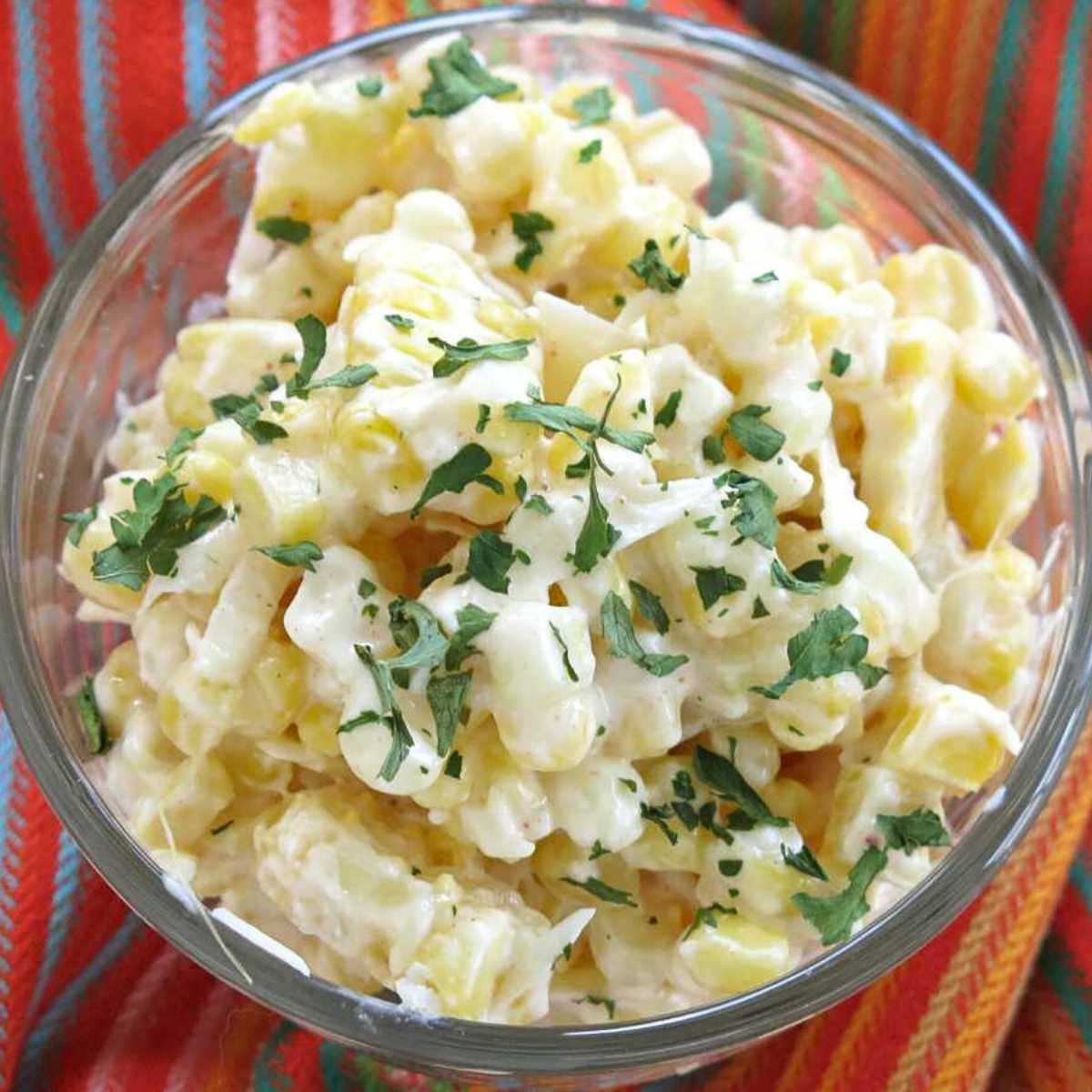 Mexican street corn in a glass cup topped with Cotija cheese and chopped cilantro on a colorful napkin.