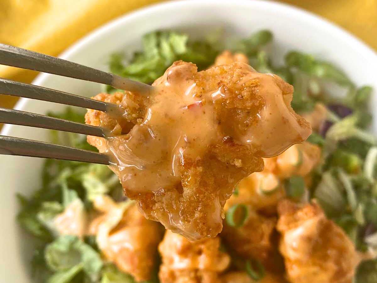 Fork holding bang bang chicken over a plate with leafy greens and more chicken.