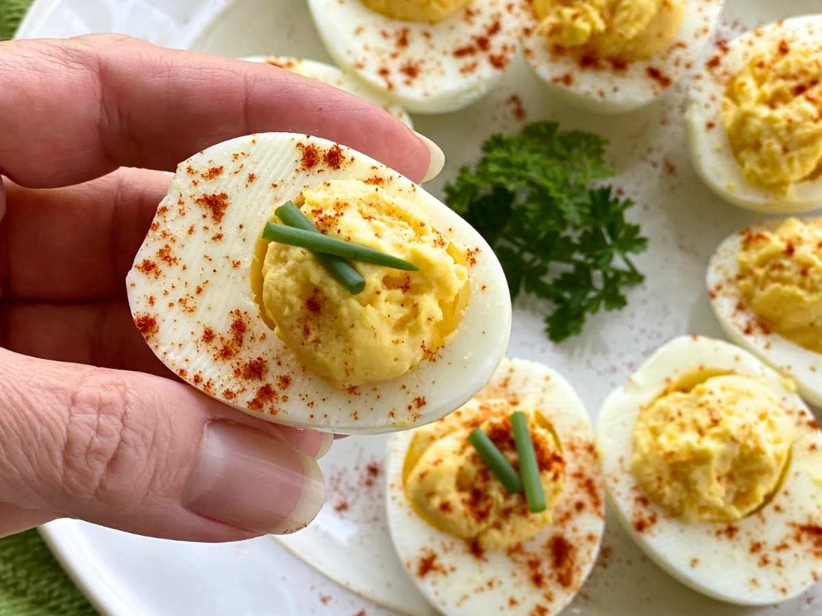 Hand holding a deviled egg over a white plate with more deviled eggs and parsley.