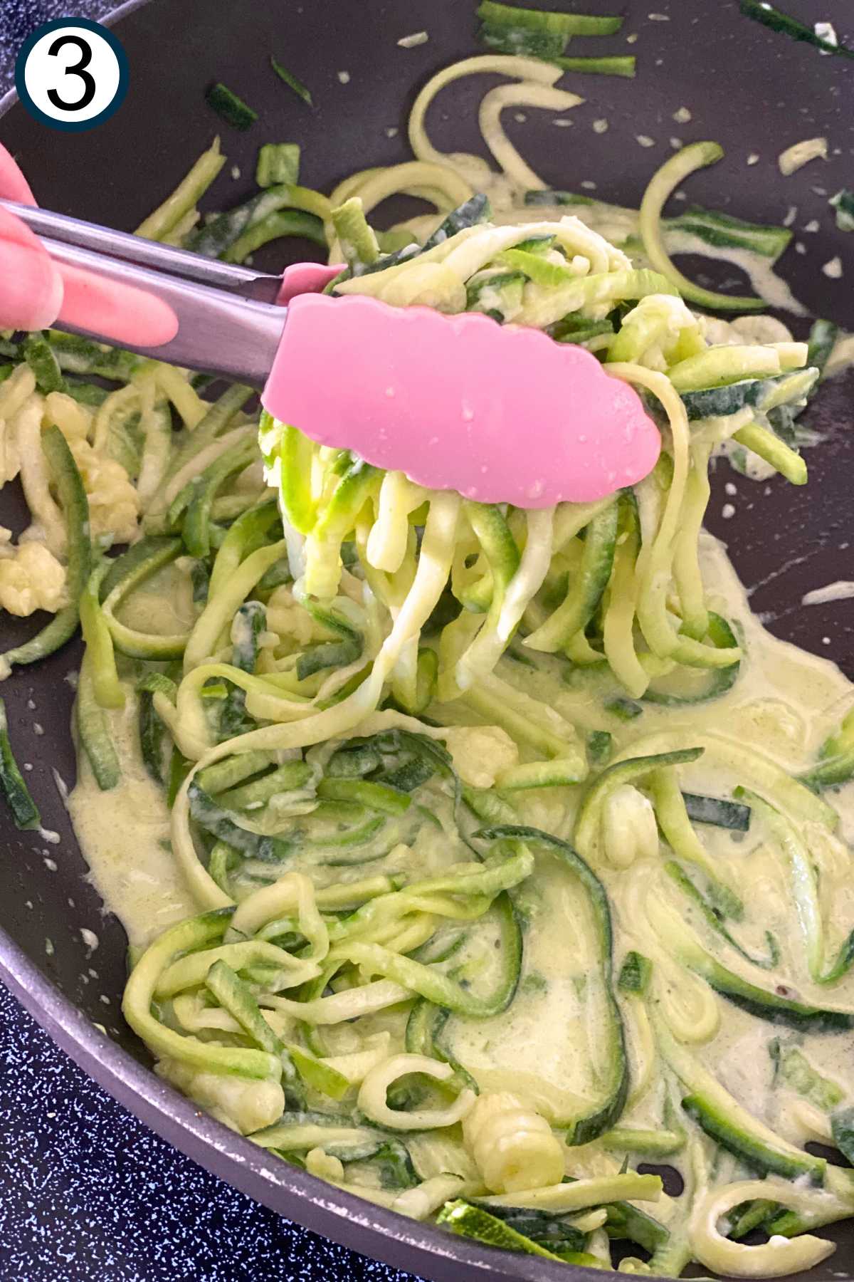 Tongs lifting zucchini noodle Alfredo out of the skillet, leaving extra sauce in the pan.
