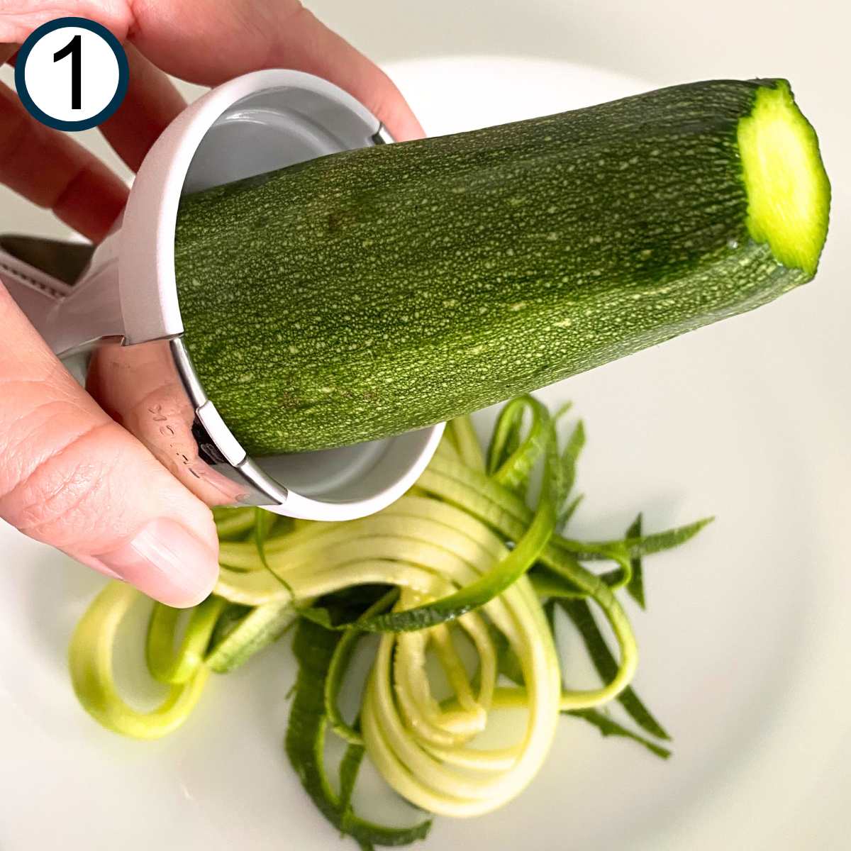 Hand holding a Veggetti spiralizer with a zucchini in it above a plate with spiralized zucchini on it.