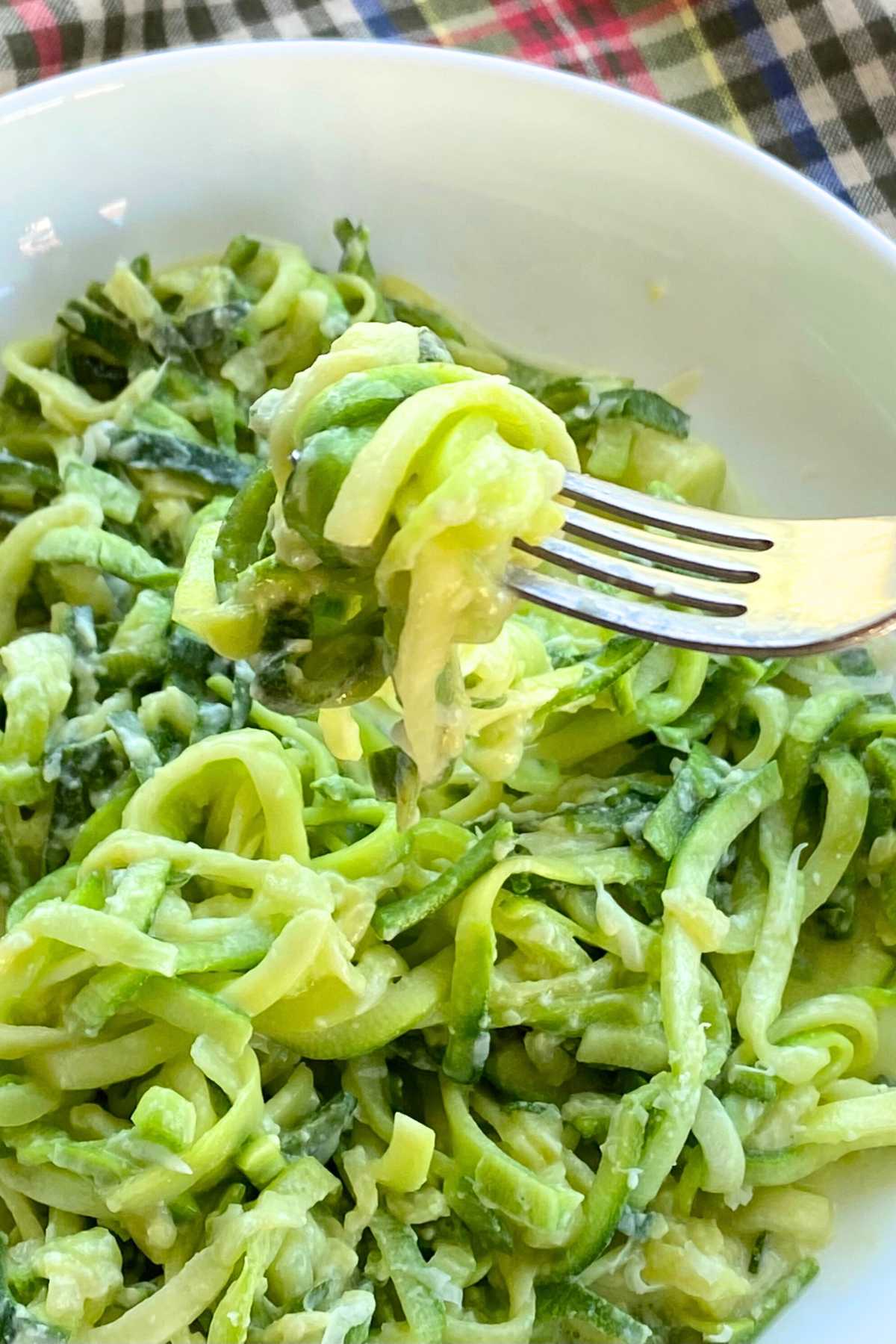 Fork lifting cooked zucchini alfredo out of a white bowl.