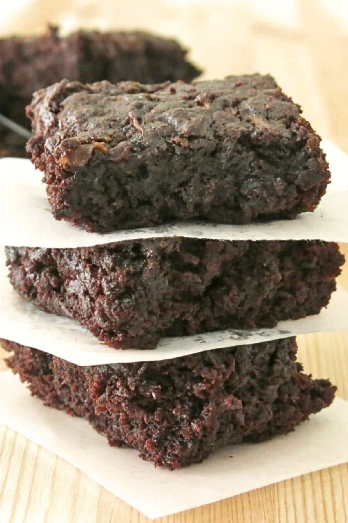 Stack of three fudgy zucchini brownies separated by parchment paper on a butcher block table top.