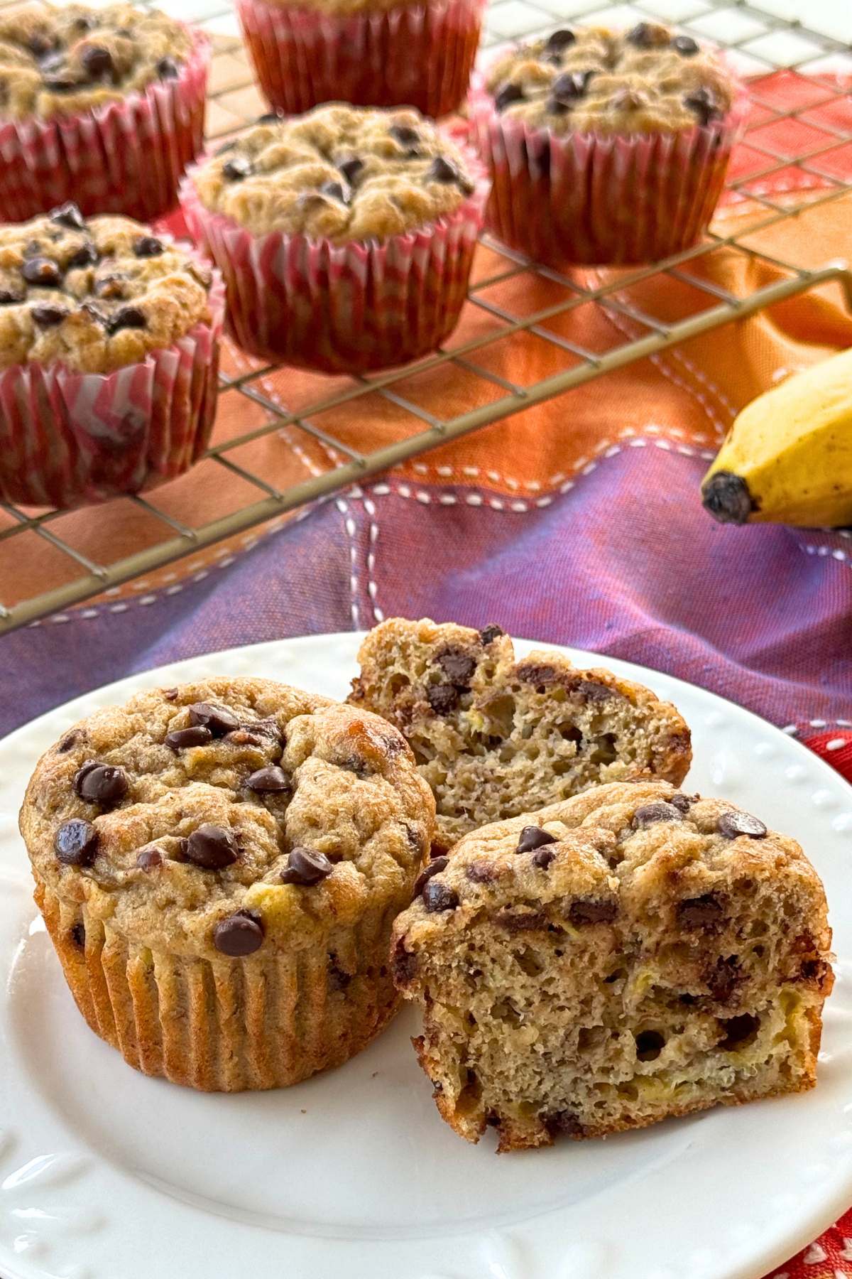Two healthy banana muffins with chocolate chips on a white plate, one is cut in half, with more on a cooling rack behind it.
