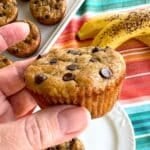 Hand holding a Greek yogurt banana muffin with two ripe bananas and more muffins behind it.