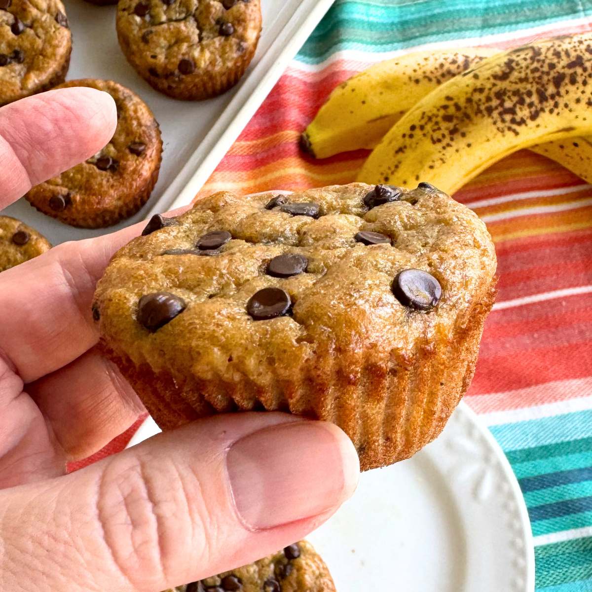 Hand holding a healthy banana yogurt muffin with ripe bananas and more muffins on a platter behind it.