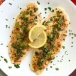 Two lemon baked basa fillets on a white plate scattered with parsley, with two lemon slices on top.
