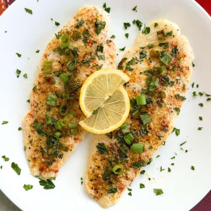 Two lemon baked basa fillets on a white plate scattered with parsley, with two lemon slices on top.