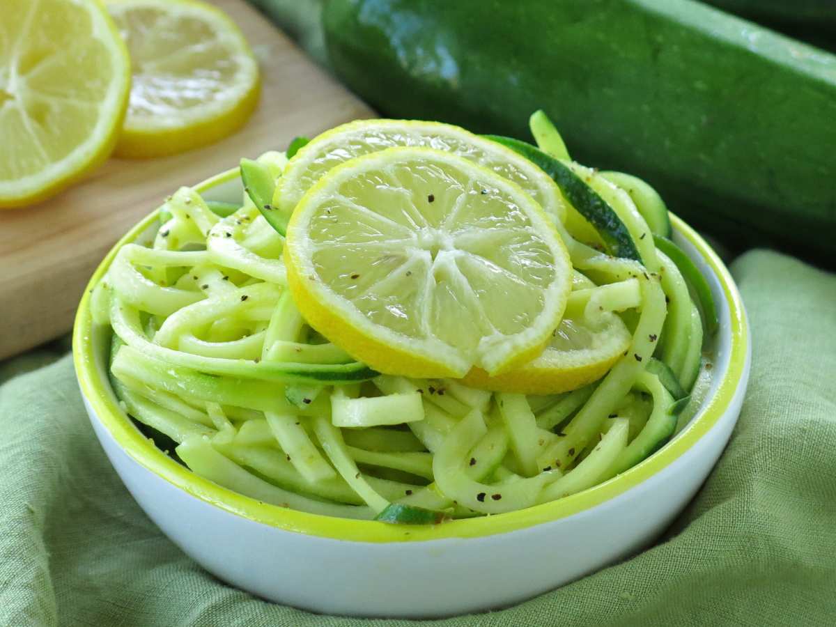 Lemon zucchini noodles in a bowl topped with slices of lemon with two zucchini gourds behind it.