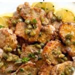 Pork piccata with a a lemon caper sauce on a white plate surrounded by lemon slices.