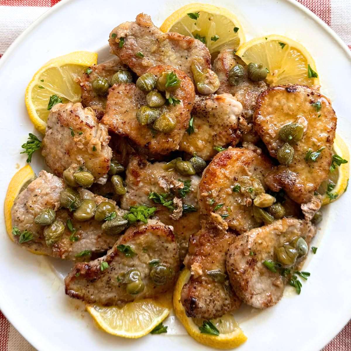 Pork tenderloin piccata with a a lemon caper sauce on a white plate surrounded by lemon slices.