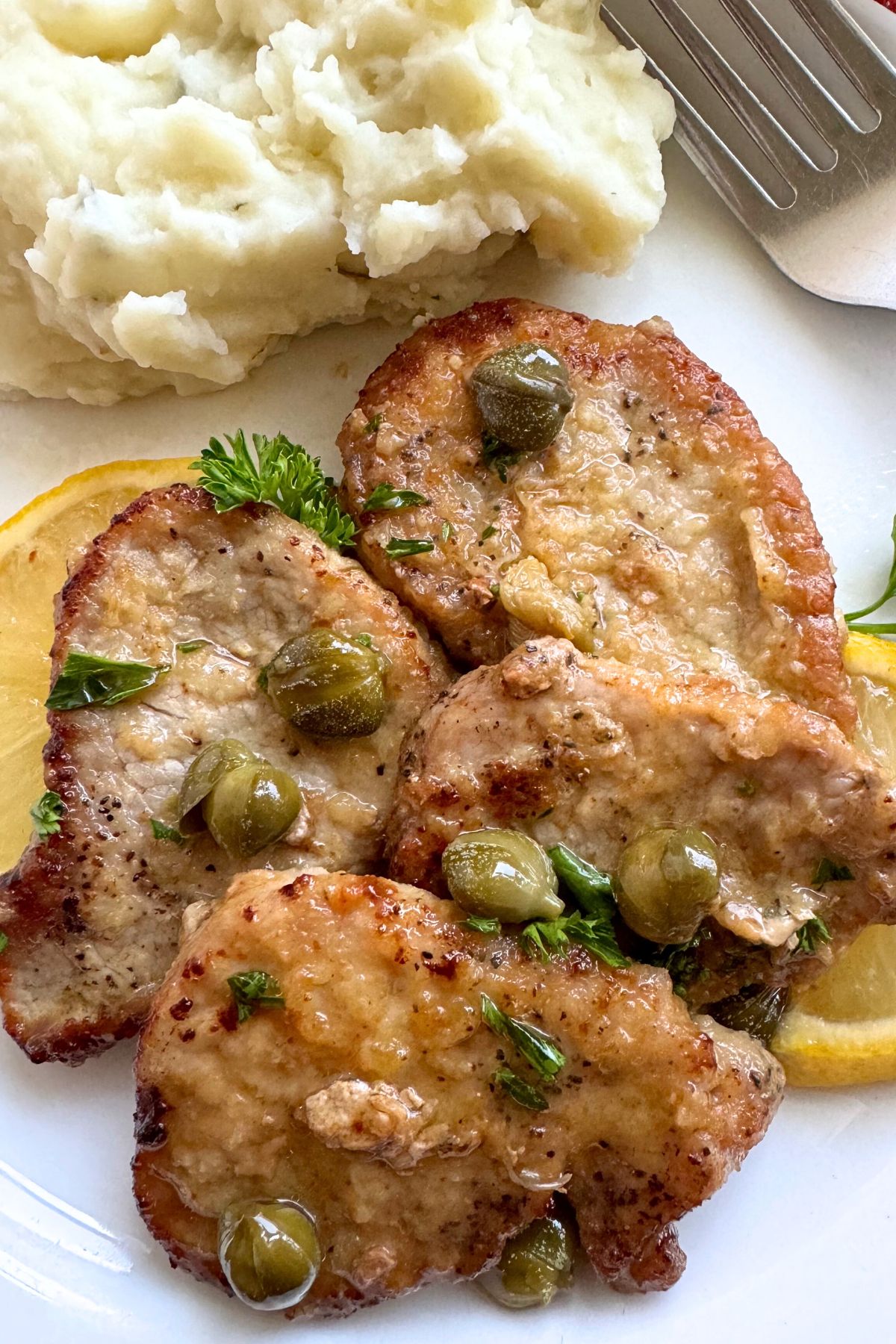Four pork tenderloin piccata medallions topped with lemon butter sauce, capers, and fresh parsley on a plate with mashed potatoes and a fork in the background.