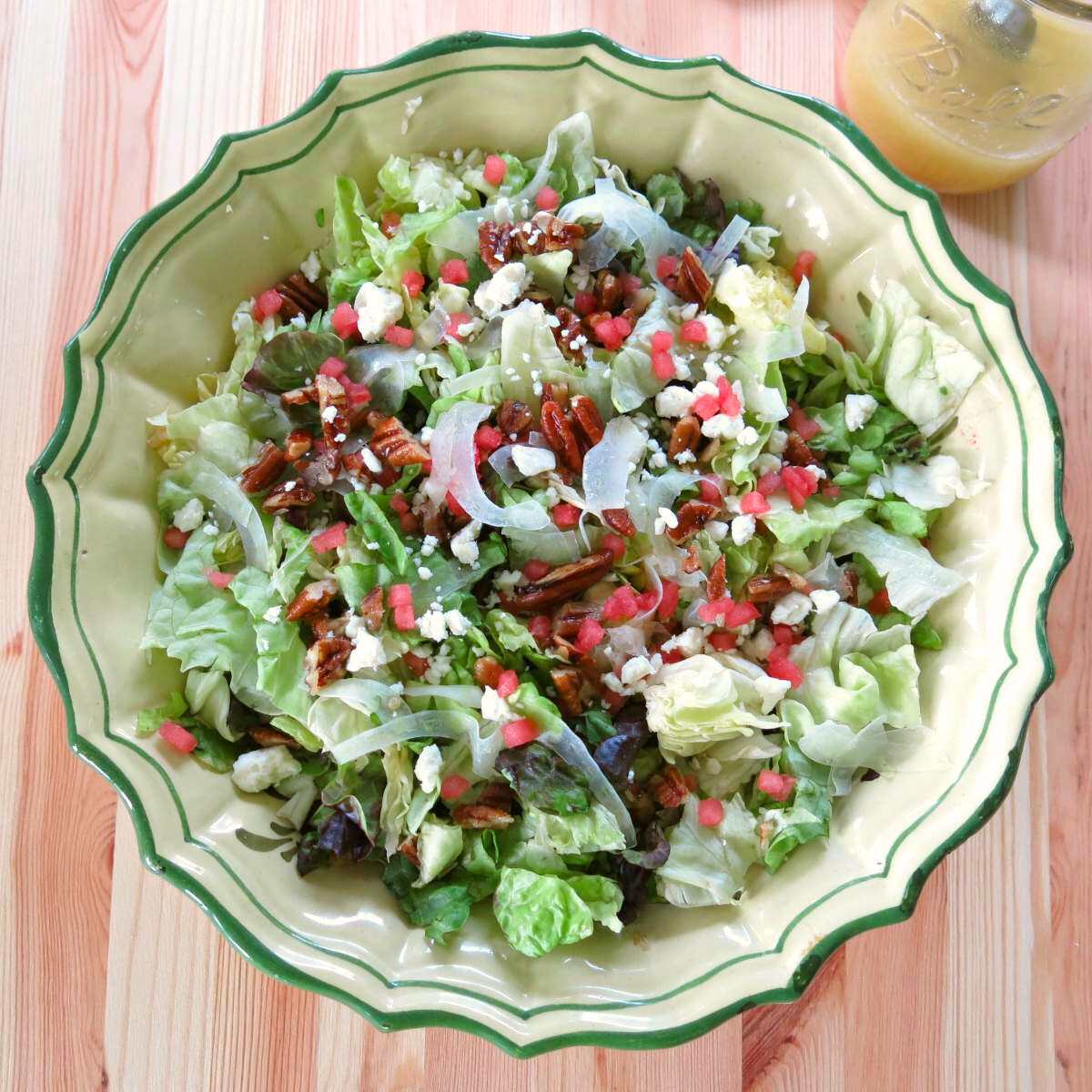 Bowl of summer salad with fennel, watermelon, feta, and candied pecans with a jar of Orange honey mustard vinaigrette in a mason jar behind it.