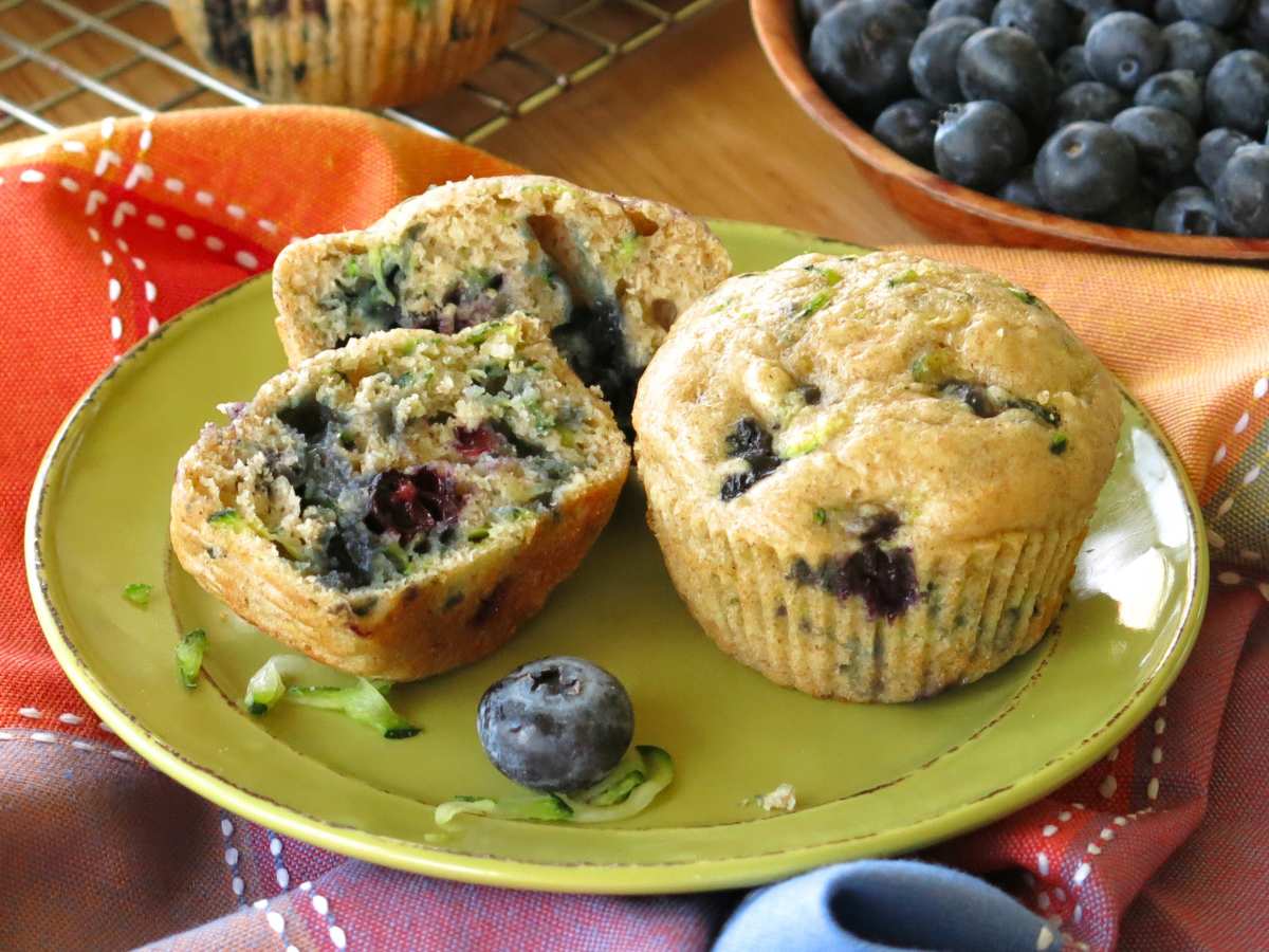 Two zucchini blueberry yogurt muffins on a green plate, one cut in half, with blueberries in the background in a bowl.