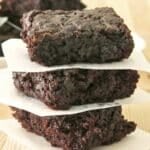 Stack of three fudgy zucchini brownies separated by parchment paper on a butcher block table top.