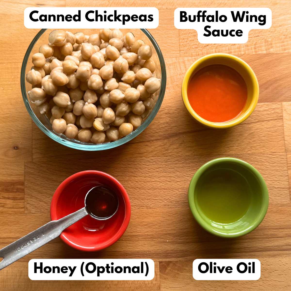 Ingredients to make the recipe, each in a separate bow: canned chickpeas, buffalo wing sauce, olive oil, and honey on a measuring spoon.