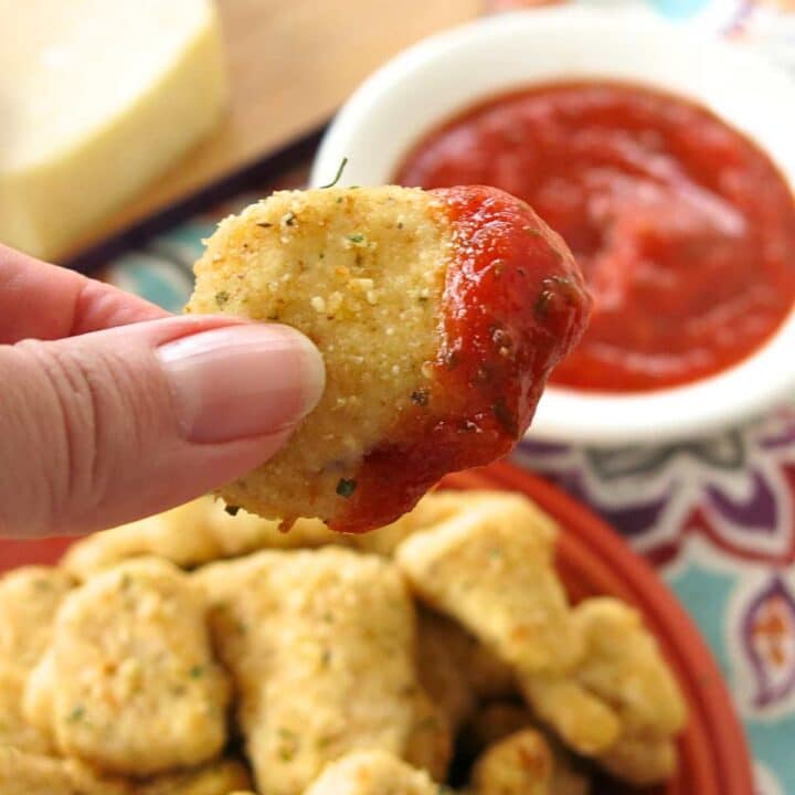 Fingers holding a garlic parmesan chicken bite that has been dipped in marinara sauce over a plate with more.